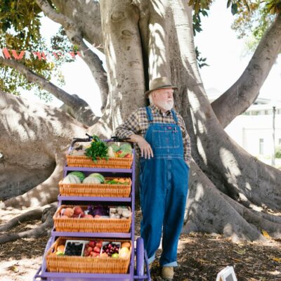 Senior farmer stands by produce cart under a large tree in a park.