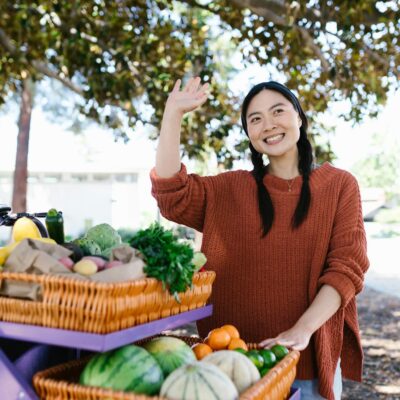 Smiling vendor standing by fruit cart waving at farmers market.