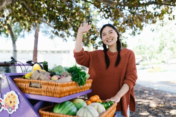 Smiling vendor standing by fruit cart waving at farmers market.